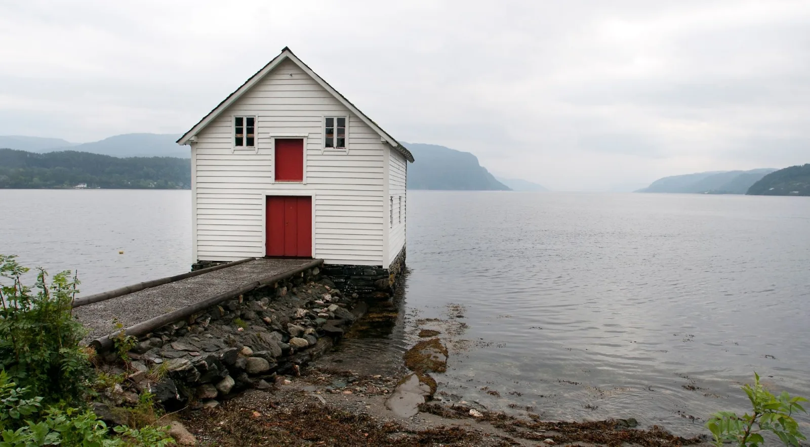 A lone white boathouse with a red door on a still, misty Lofoten fjord